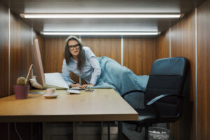 Stressed, frustrated employee wakes up in a cubicle cot, installed by her employer to improve work-life balance and productivity.