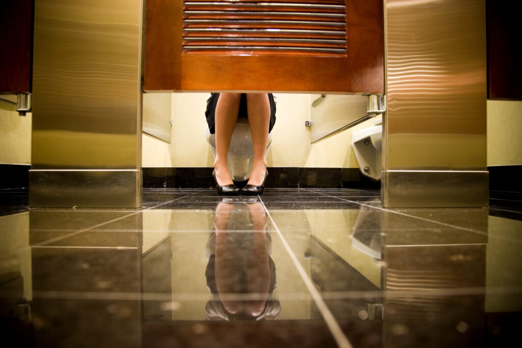 A stressed office worker crying in a fluorescent-lit bathroom stall, symbolizing corporate burnout and absurd wellness culture.